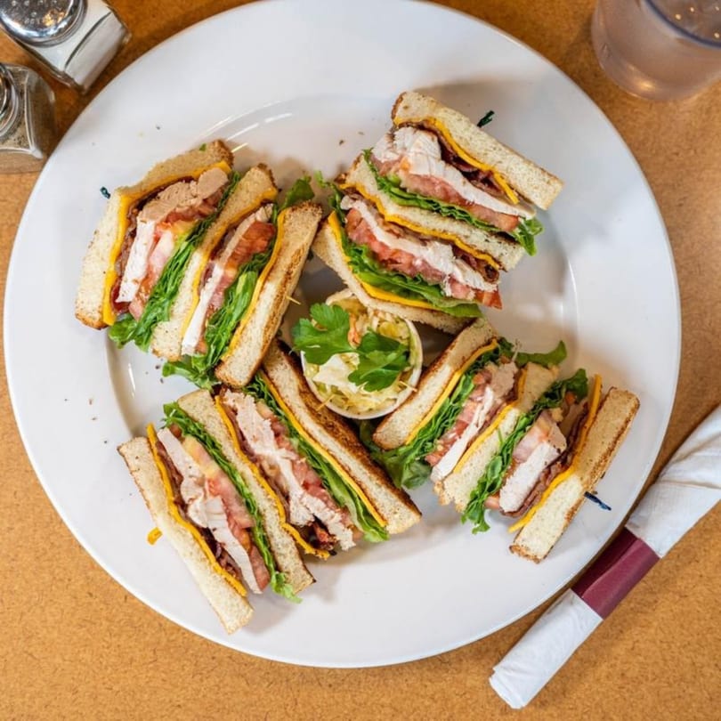 A Stella's Diner club sandwich, cut into quarters, with a side of coleslaw on a white plate.