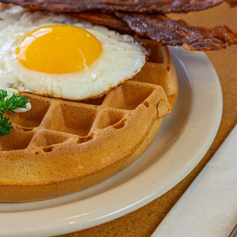A Stella's Diner waffle topped with a fried egg and crispy bacon on a white plate.