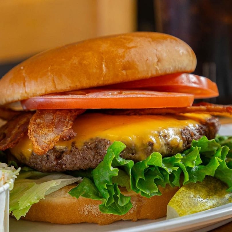 A Stella's Diner bacon cheeseburger with lettuce, tomato, and a pickle spear on a white plate.
