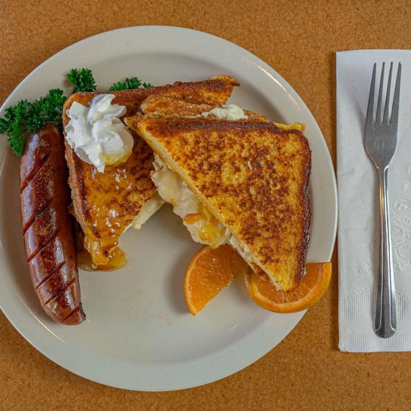 French toast with whipped cream, syrup, sausage, and orange slices on a white plate.