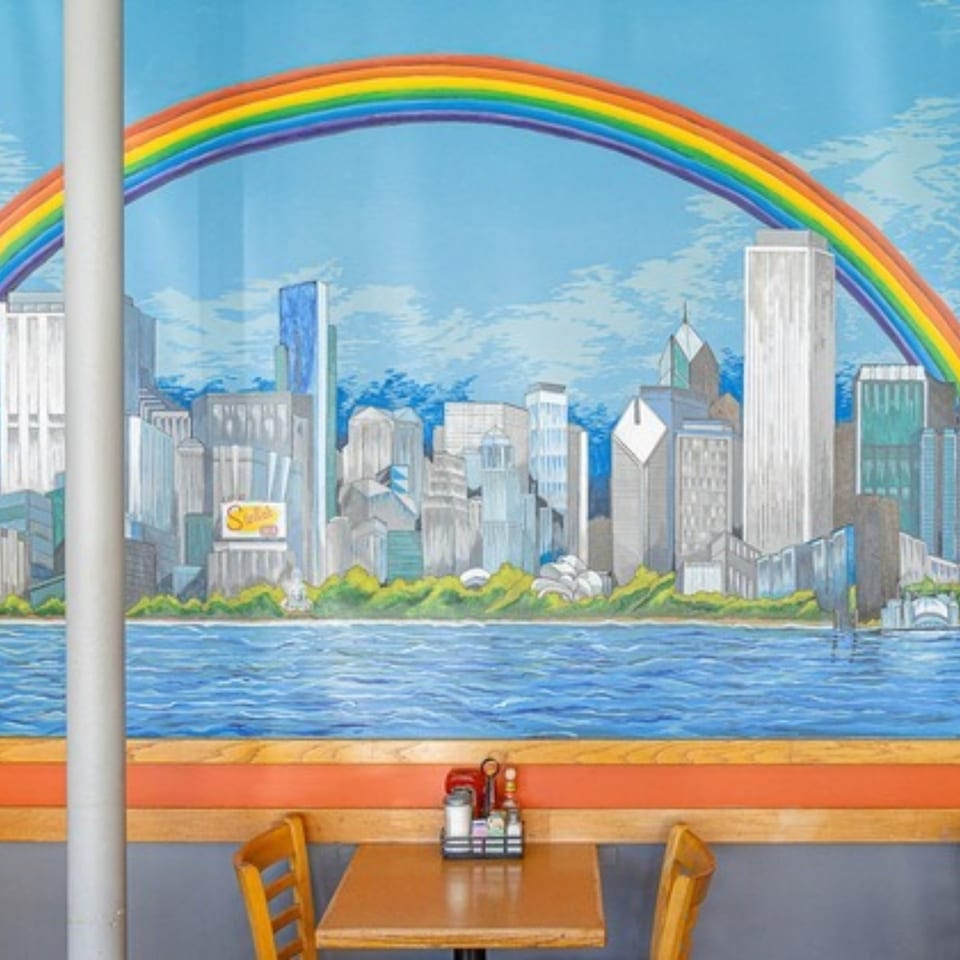 A vibrant mural of a city skyline with a rainbow over water, above a diner table setting.